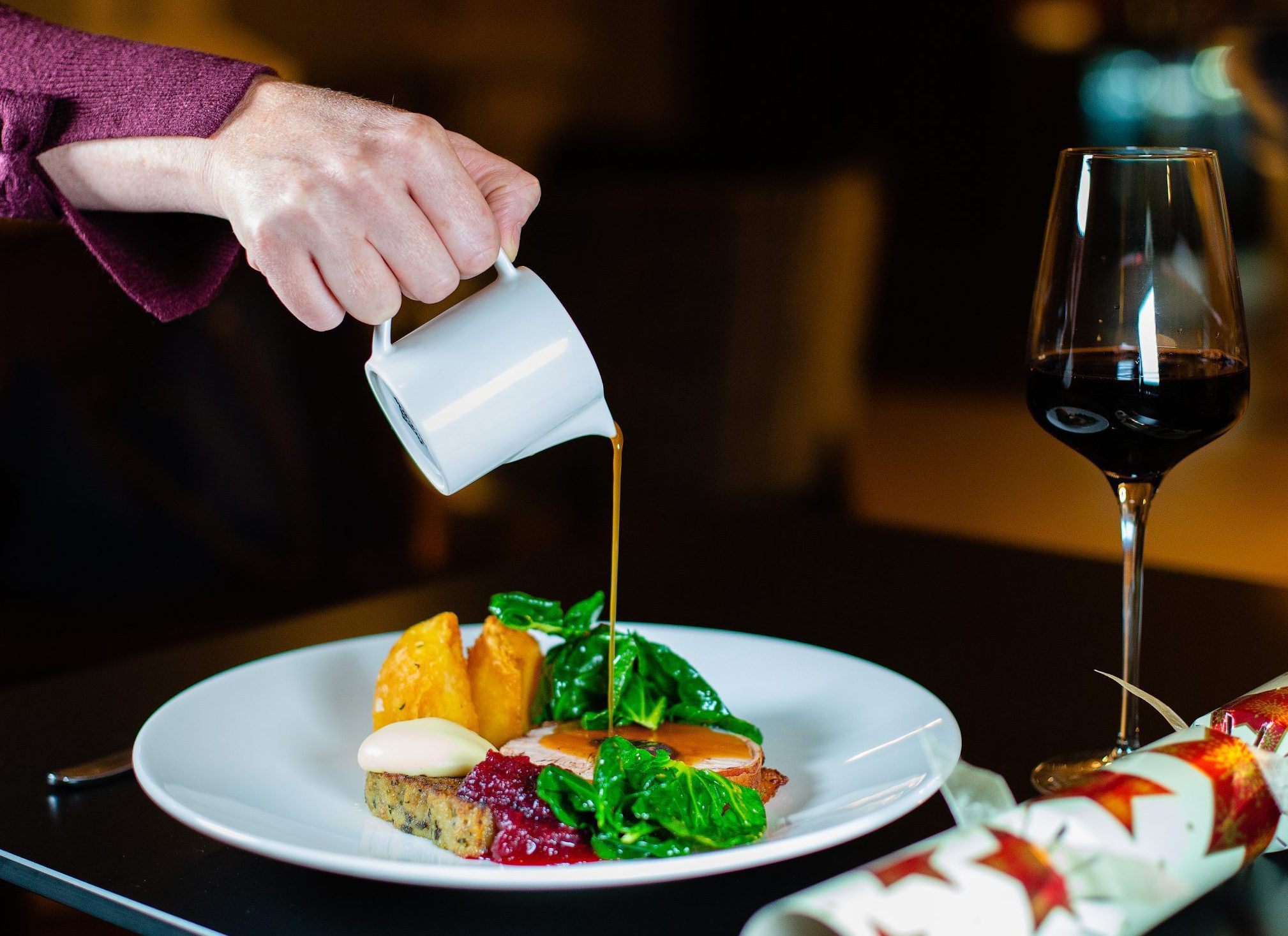 Gravy being poured onto Christmas Dinner next to a glass of red wine and a Christmas cracker