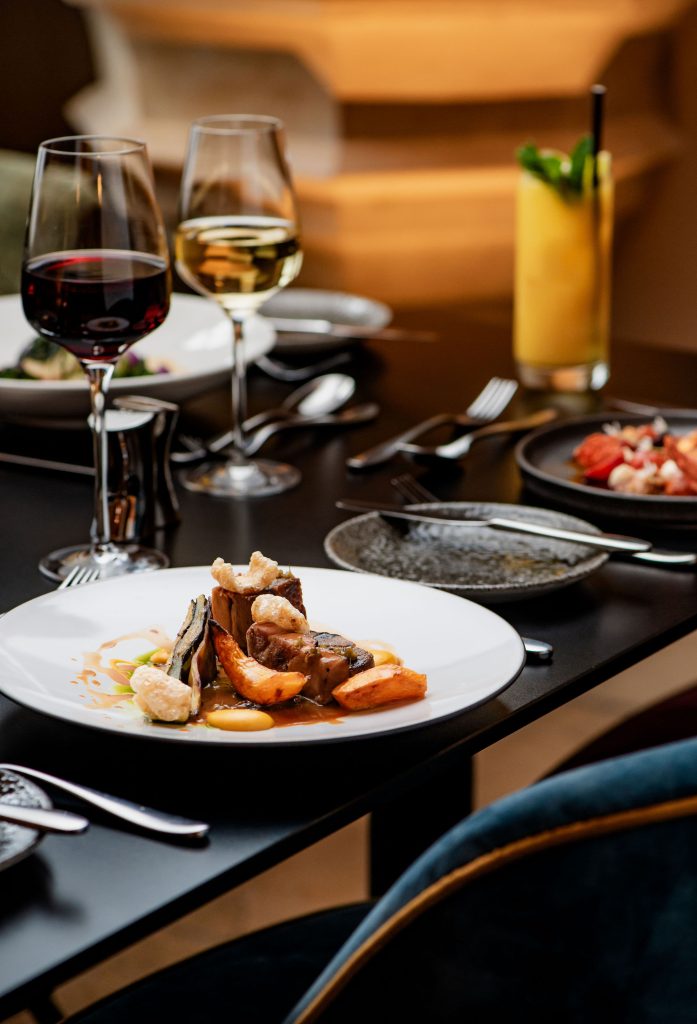 A table of food and wine. The table has red wine and wgute with a main course of Slow Cooked Large Black Pig Belly & Grilled Jowl, Sand Carrots, Pancetta Jam, Wild Garlic and Potato Galette Blanquette Sauce