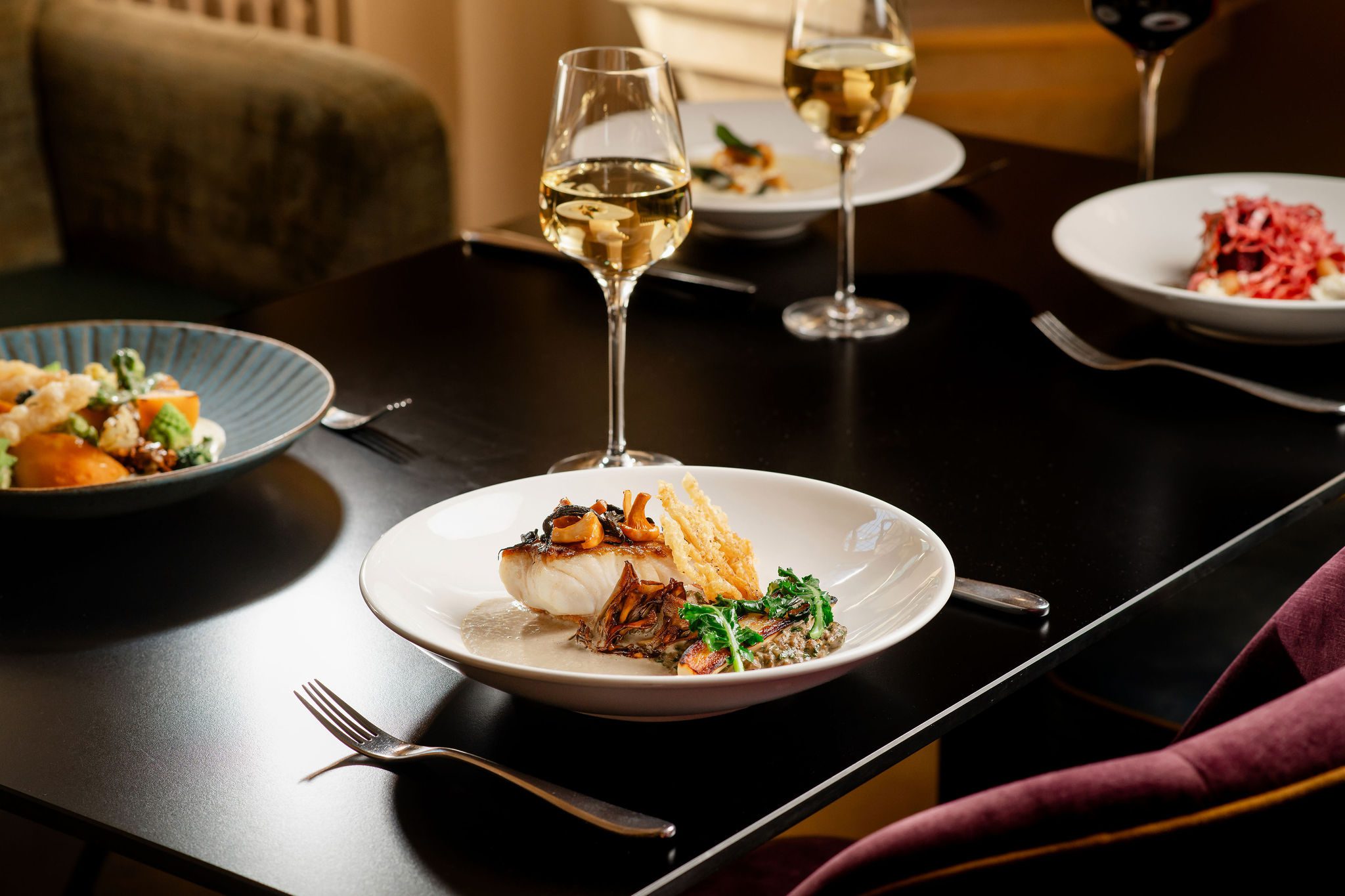 A photo showcasing the food served in the MCR. The table focuses on a plate of food with a white wine - South Coast Cod , Pearl Barley, Salsify, Black Trompetes, BBQ Maitake and a Sauce of Roasted Yeast