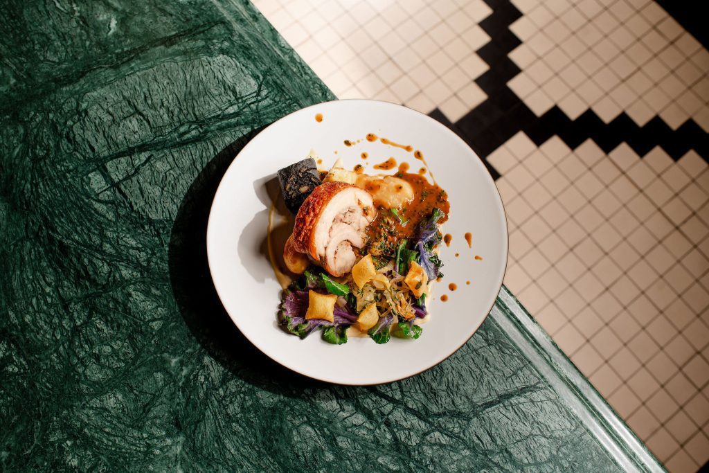 A photo showcasing the food served in the MCR. The table focuses on a plate of food - Old Spot Pork belly, Black Pudding, Choucroute Garni, Wild Cabbages, Apple and Café de Paris Sauce